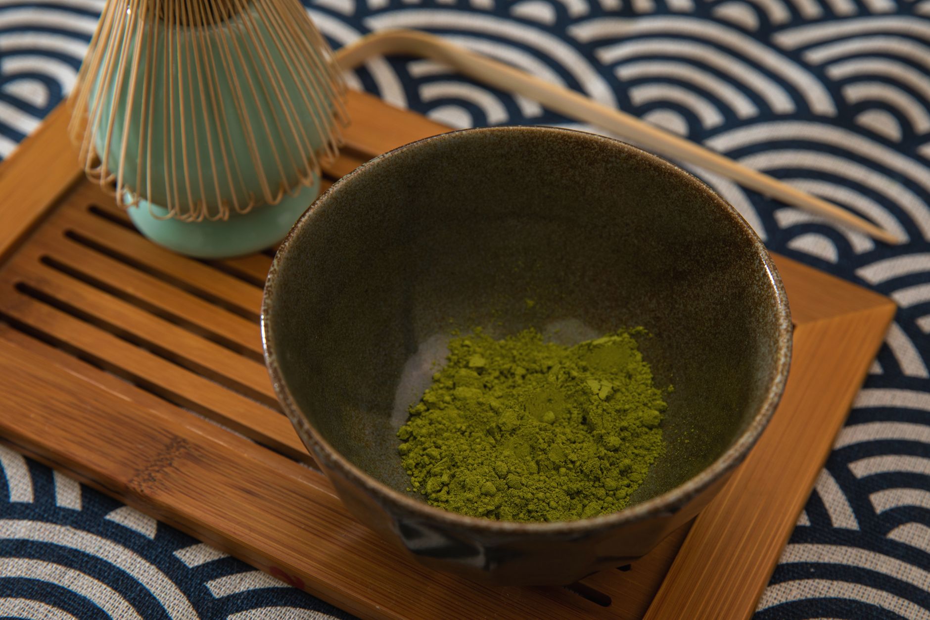 Bol en céramique contenant de la poudre de matcha sur un plateau en bambou, avec un fouet à thé et une cuillère en bambou sur un tissu à vagues.