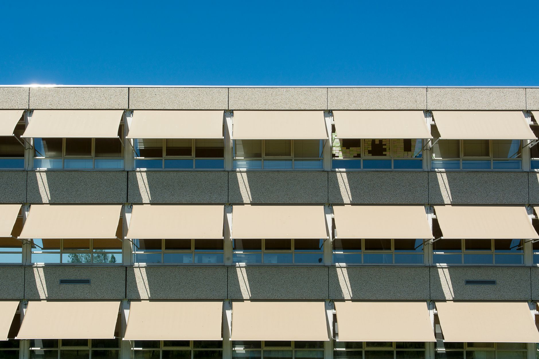 Façade d’un bâtiment en béton dont les fenêtres en bandeau sont protégées par des stores bannes beiges déployés, sous un ciel bleu.
