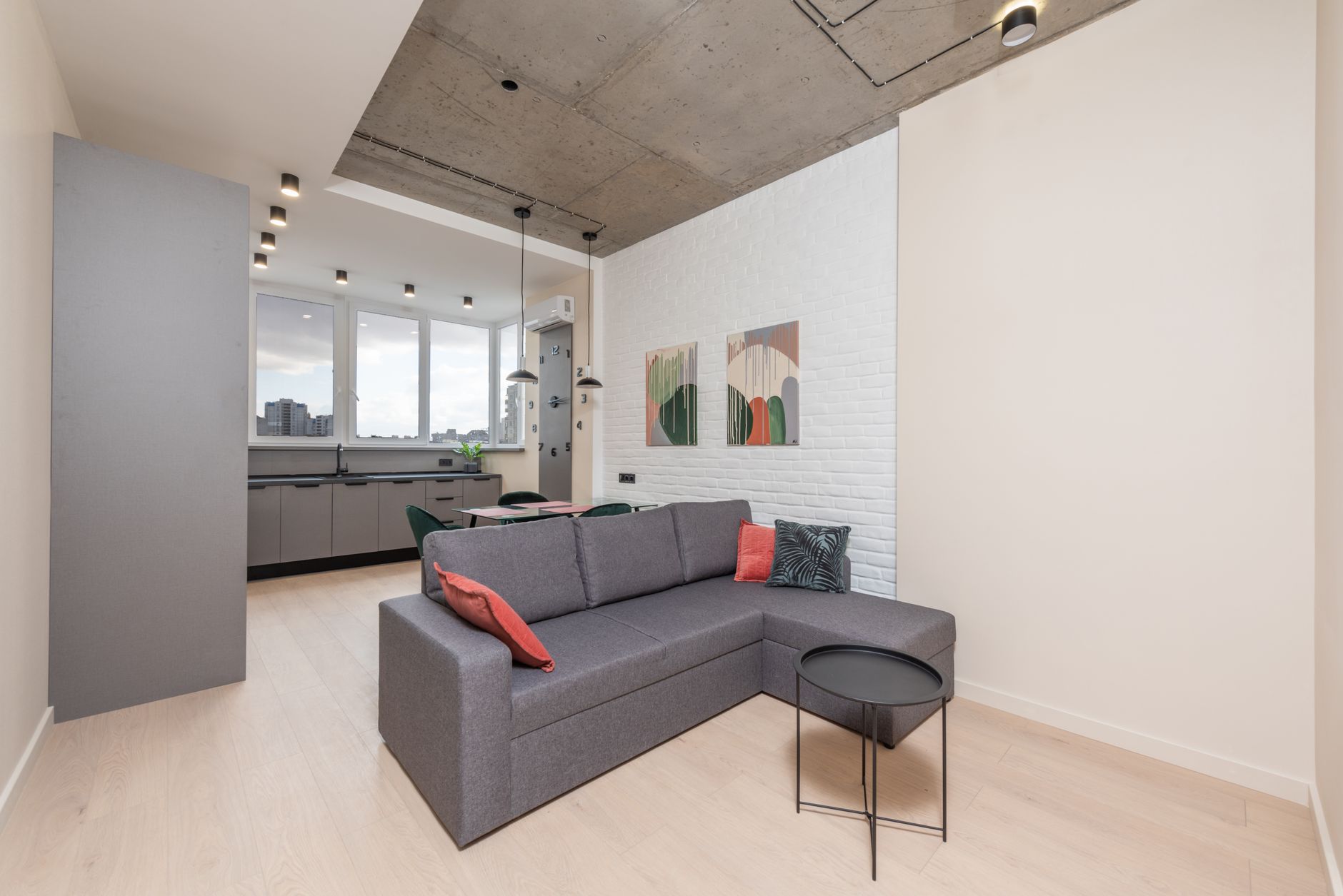 Séjour moderne avec canapé d’angle gris, mur de briques blanches, plafond en béton et cuisine ouverte au fond, sous des spots.