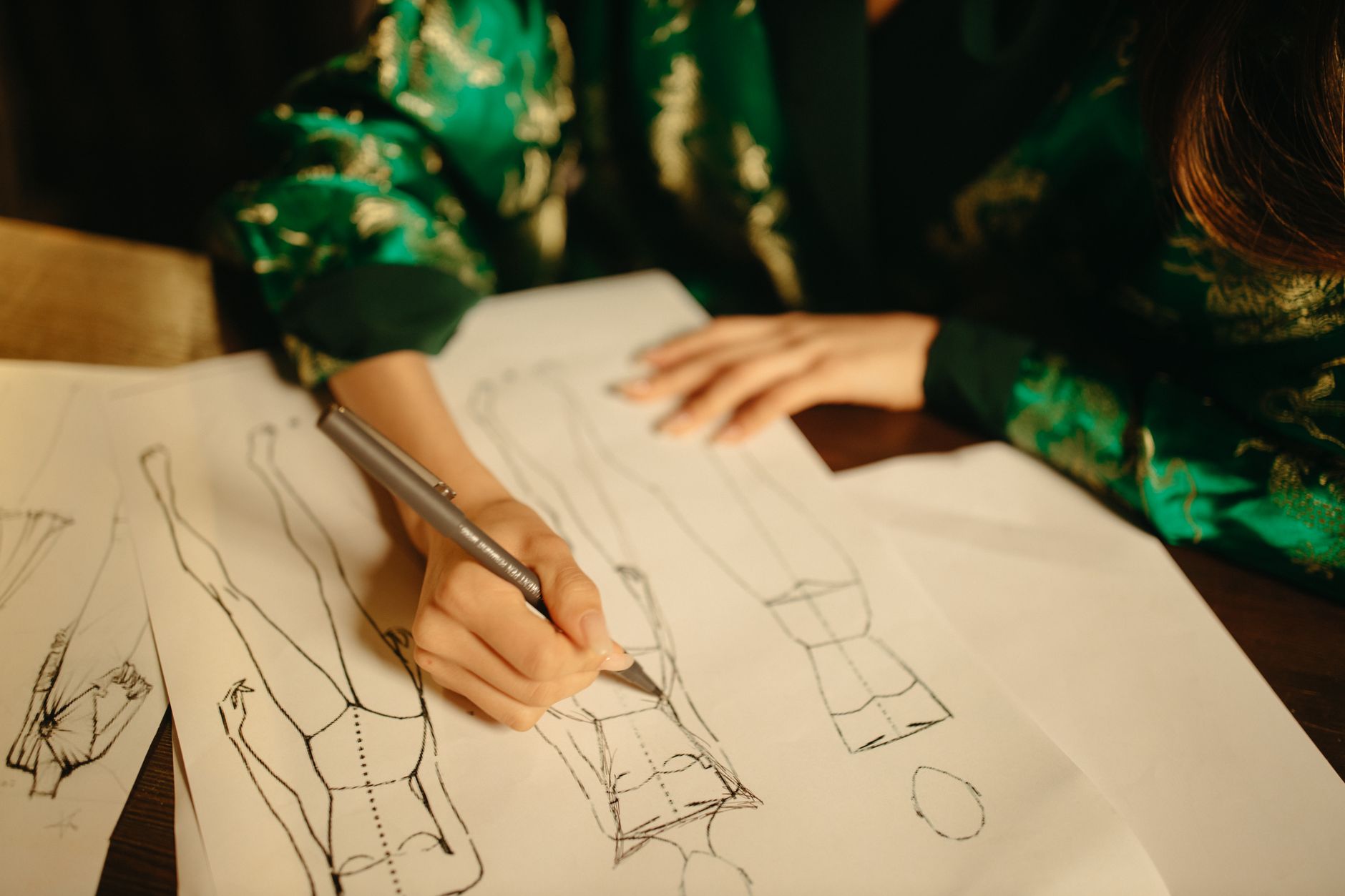 Une personne en tenue verte à motifs dorés dessine au stylo des silhouettes de mode sur des feuilles posées sur une table en bois.