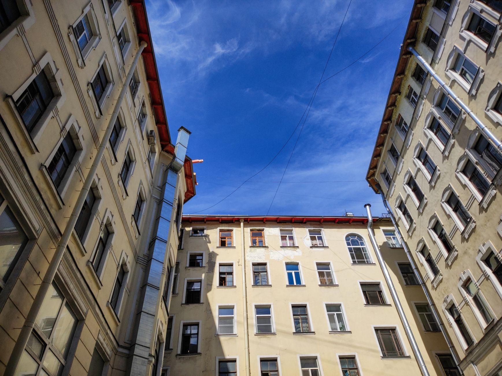 Vue vers le ciel depuis une cour intérieure entourée d’immeubles d’habitation anciens aux façades claires, avec fenêtres alignées, descentes d’eau et ciel bleu.